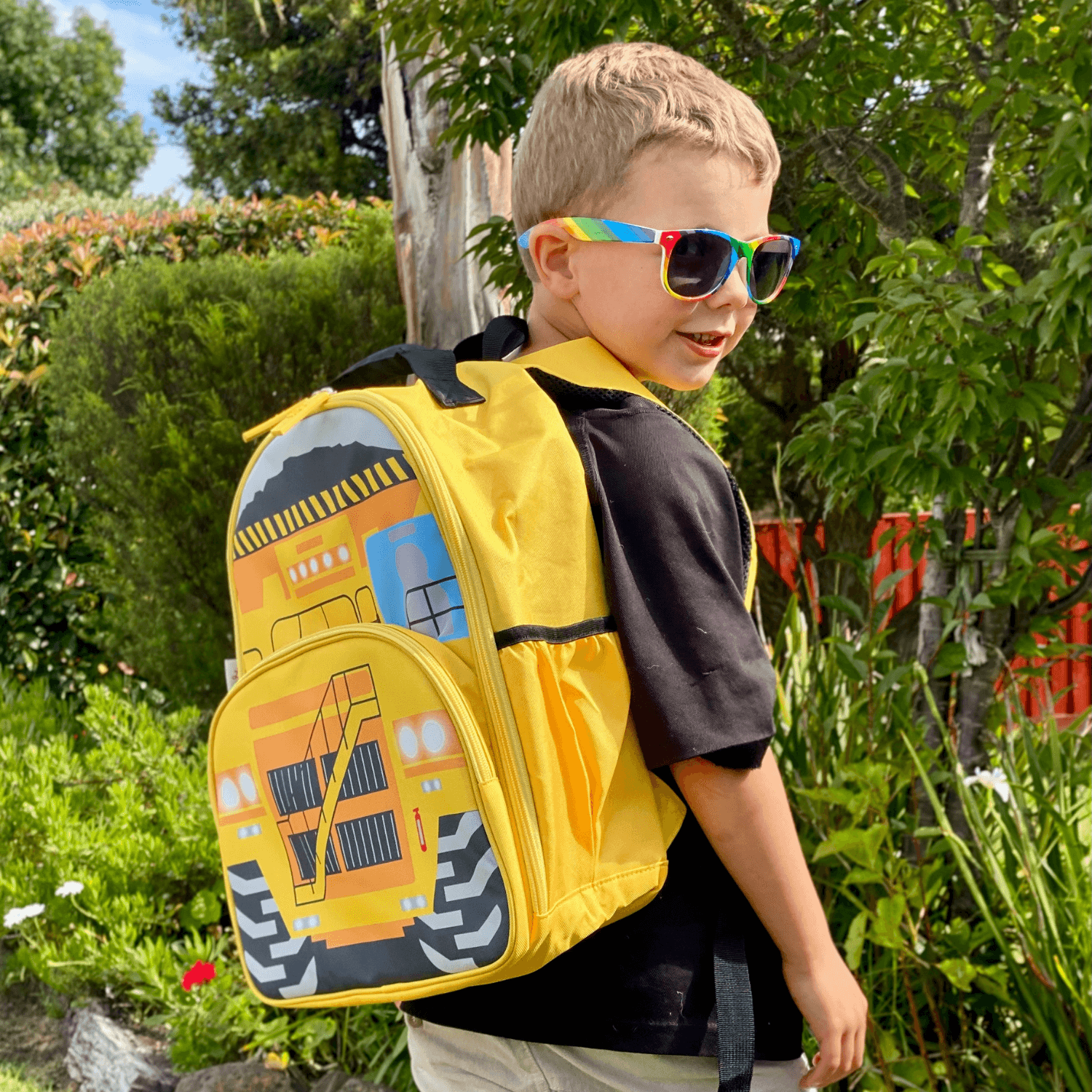 Boy wearing a jude&moo Australia Yellow Mining Dump Truck Kids Backpack in front of a garden.