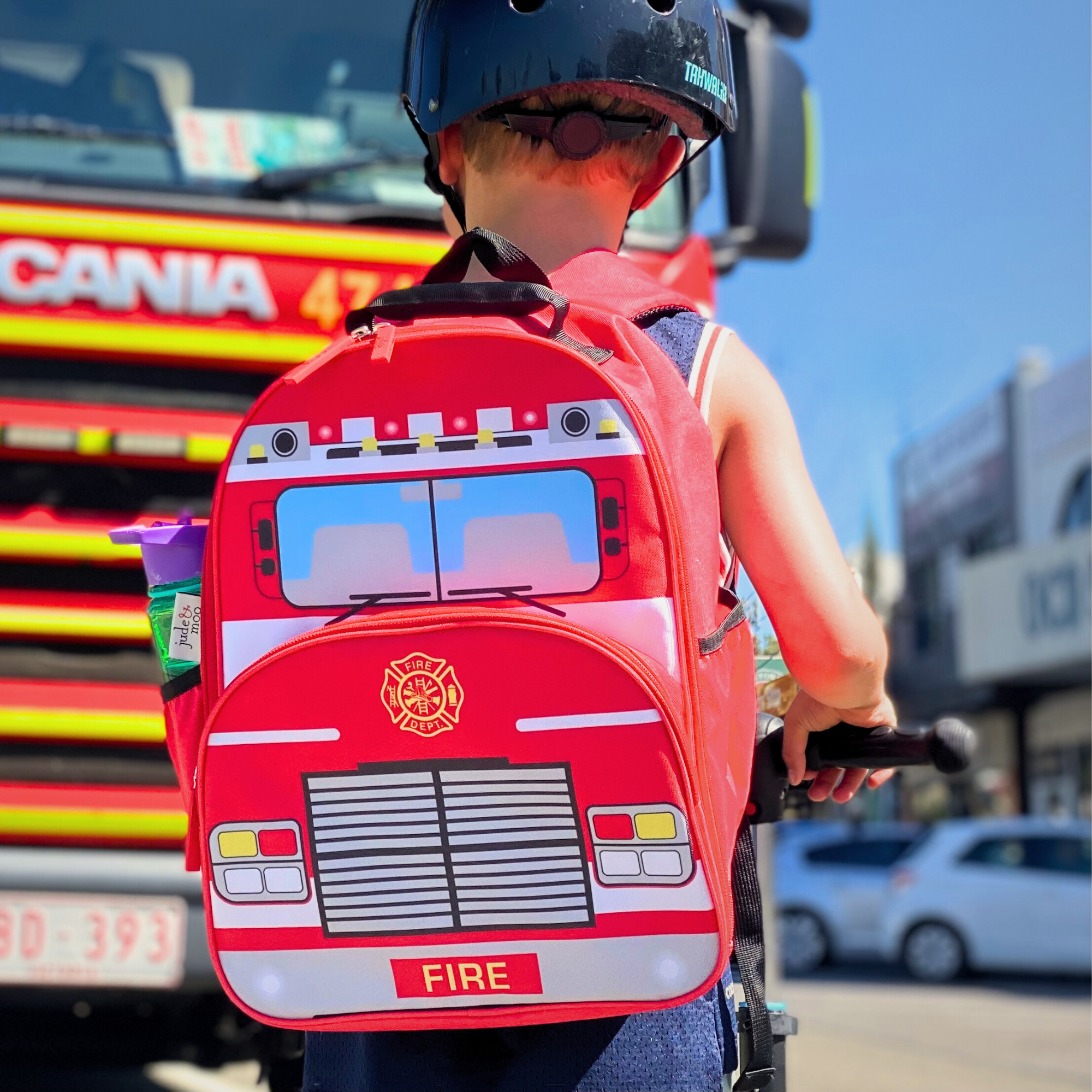 Boy standing in front of a fire engine wearing jude&moo Kinder Fire Truck Backpack 