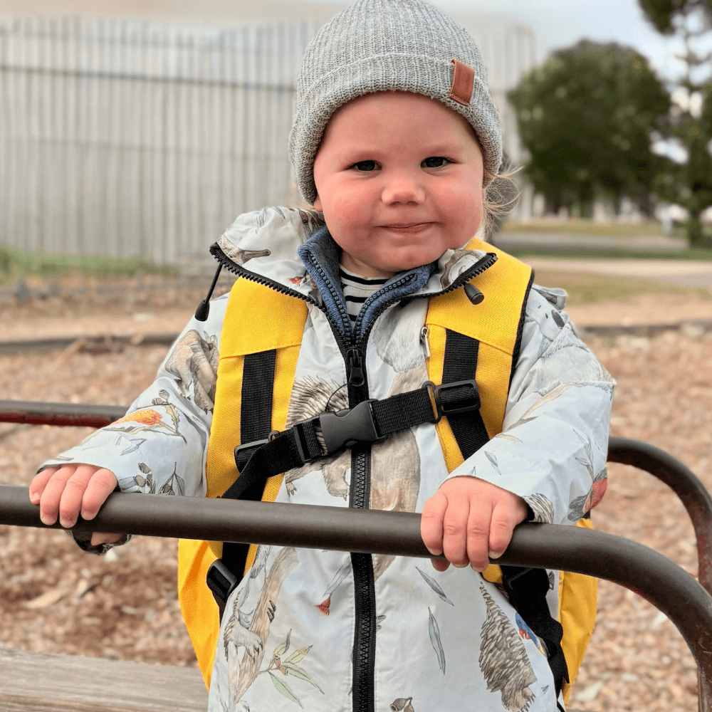 Child wearing a yellow dump truck backpack and gray beanie on a playground.